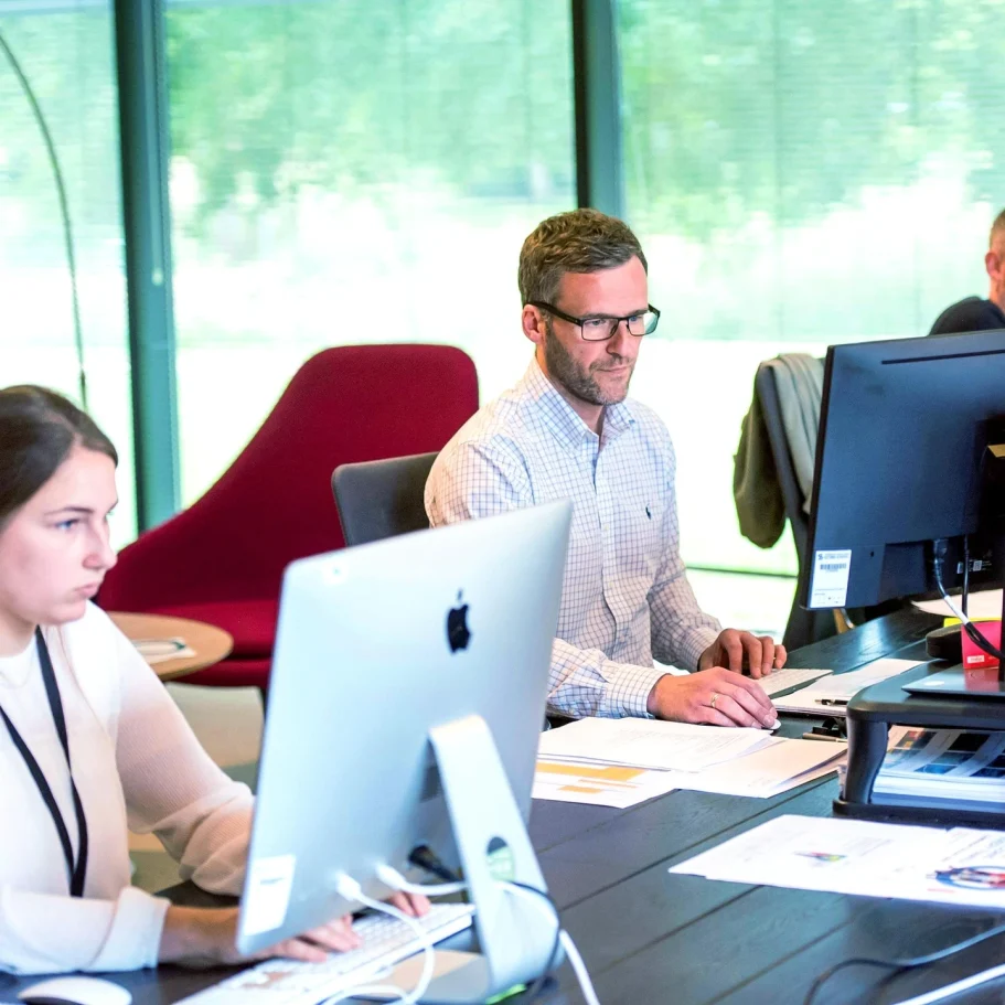 Büros & Großräume Zwei Personen arbeiten an Computern in einem modernen Büro. Ein Kollege sitzt am Tisch.